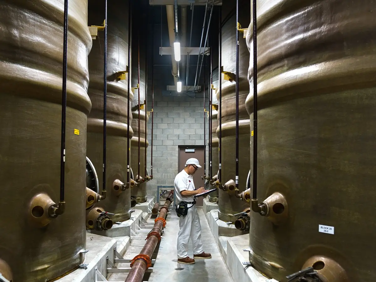 A worker in a white uniform and helmet inspects and takes notes between large industrial storage tanks in a facility.