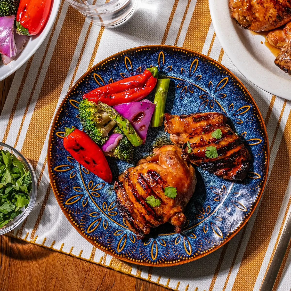 A blue plate with two grilled chicken thighs, grilled red peppers, broccoli, and purple onions on a striped tablecloth.
