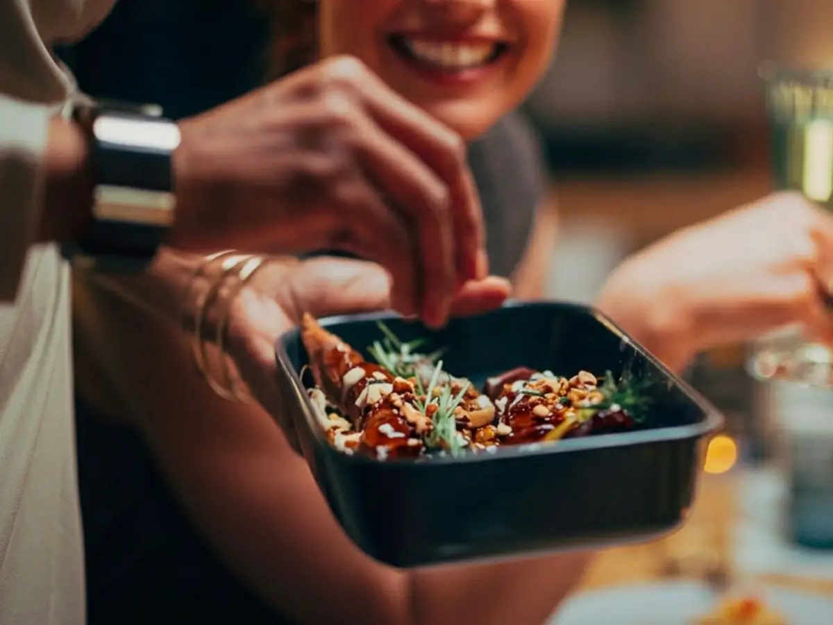 Person garnishing food in a black rectangular dish, with another person smiling in the background.