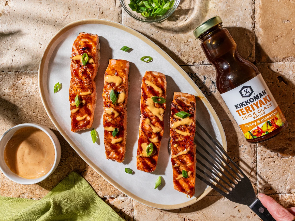Four grilled salmon fillets on a white oval plate, garnished with sliced green onions and sauce, next to a bottle of Kikkoman Teriyaki BBQ glaze and a small bowl of sauce.