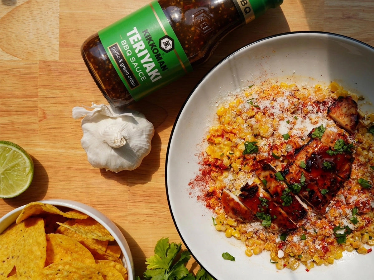 A bowl of grilled chicken with sauce on seasoned corn, next to a bottle of teriyaki BBQ sauce, garlic, lime, fresh cilantro, and a bowl of tortilla chips on a wooden surface.
