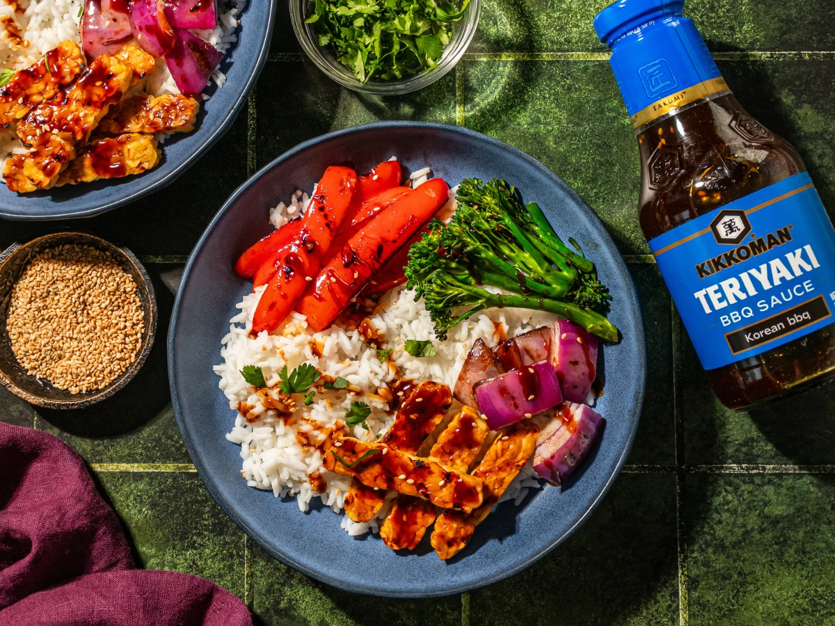A bowl with rice, grilled tempeh, carrots, broccolini, red onions, and sauce, next to a bottle of Kikkoman Teriyaki BBQ Sauce and a dish of sesame seeds on a green surface.