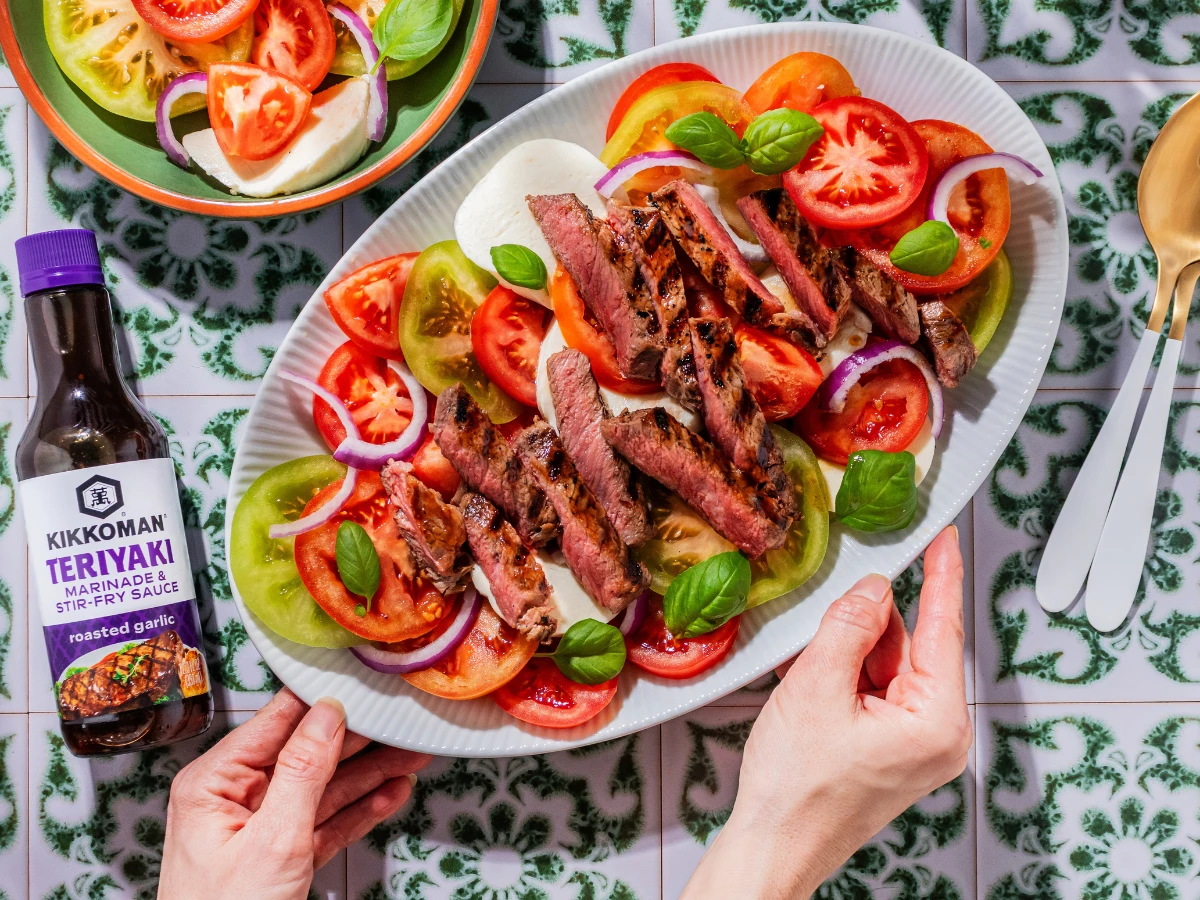 A plate of grilled steak slices with tomatoes, red onions, basil, and mozzarella, beside a bottle of Kikkoman Teriyaki sauce on a patterned tile surface.