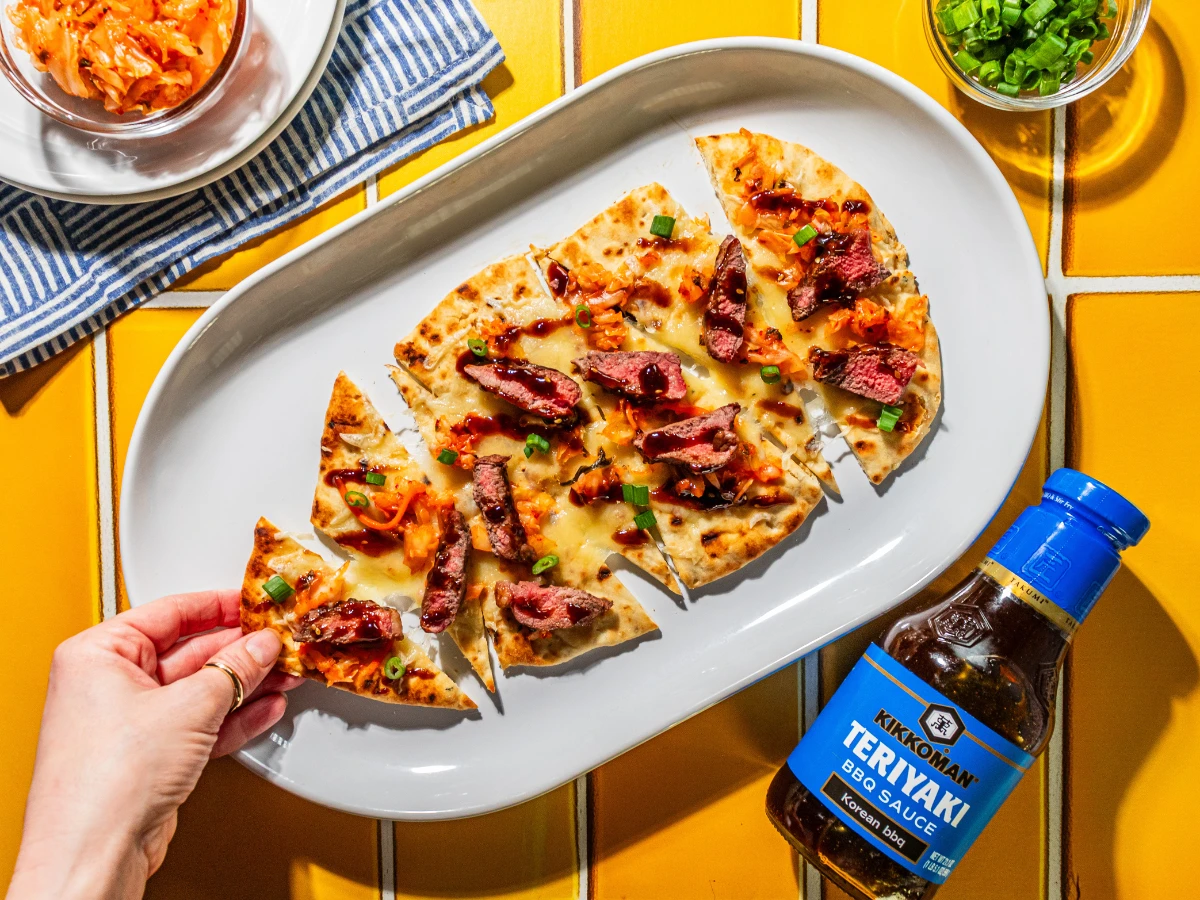 A hand takes a slice of flatbread topped with cheese, steak, green onions, and kimchi, with a bottle of Kikkoman Teriyaki BBQ Sauce and a bowl of sliced green onions on the side.
