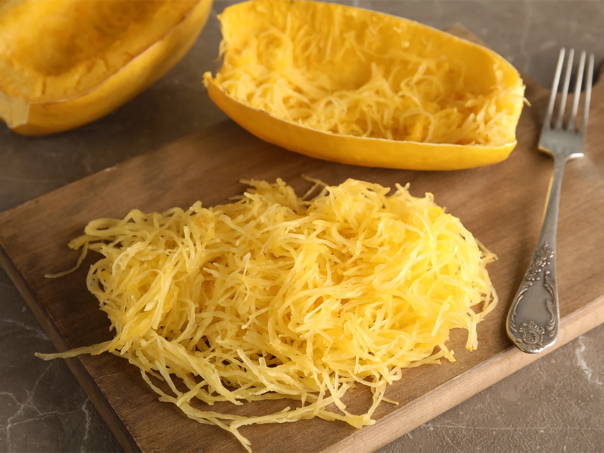 Cooked spaghetti squash flesh separated into strands on a wooden board, with empty squash shells and a fork beside it.