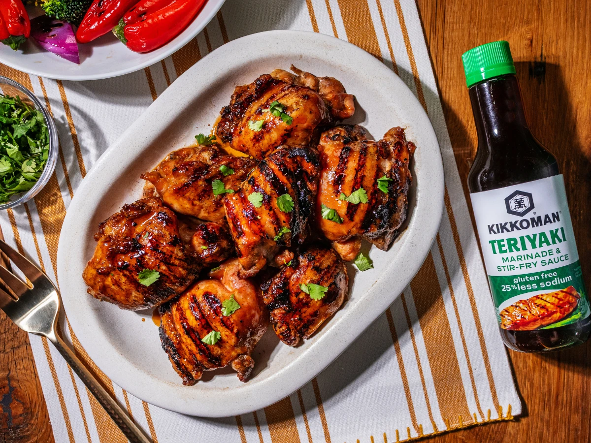 Grilled chicken thighs garnished with herbs on a white platter, next to a bottle of Kikkoman Teriyaki sauce, placed on a striped cloth with salad and herbs nearby.