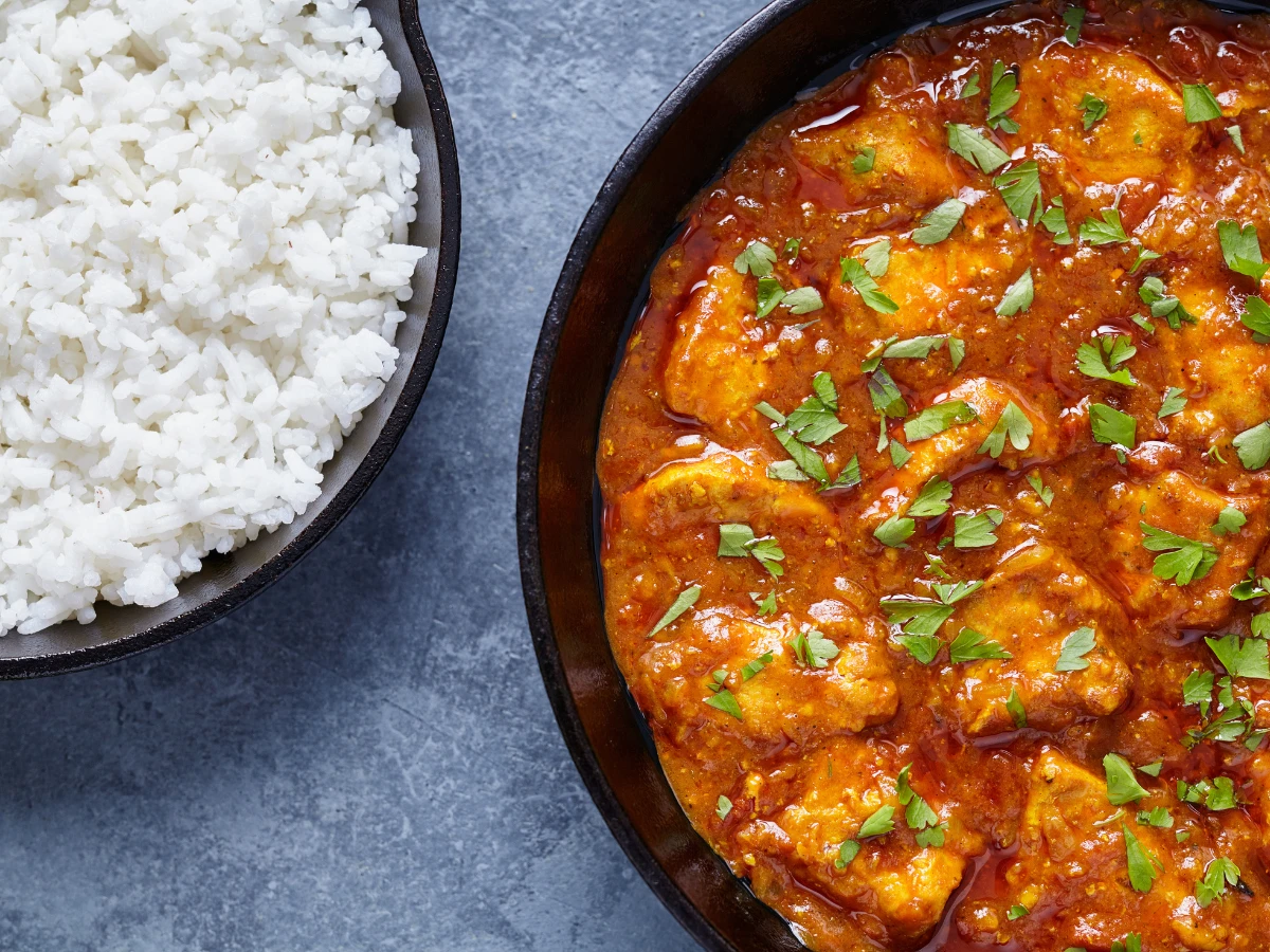 A bowl of white rice next to a pan of chicken curry garnished with chopped parsley on a gray surface.