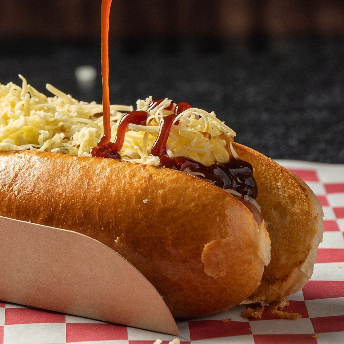 A hot dog in a paper tray topped with shredded cheese, with barbecue sauce being poured on top, set on a red and white checkered paper.
