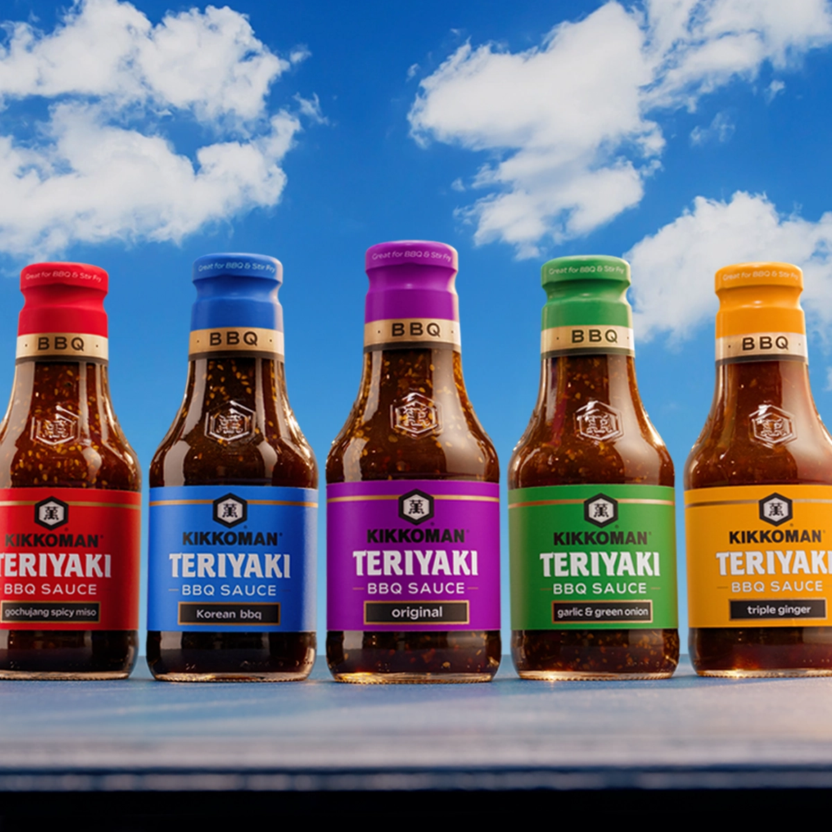 Five bottles of Kikkoman Teriyaki BBQ Sauce in different flavors are lined up on a table against a blue sky with clouds.