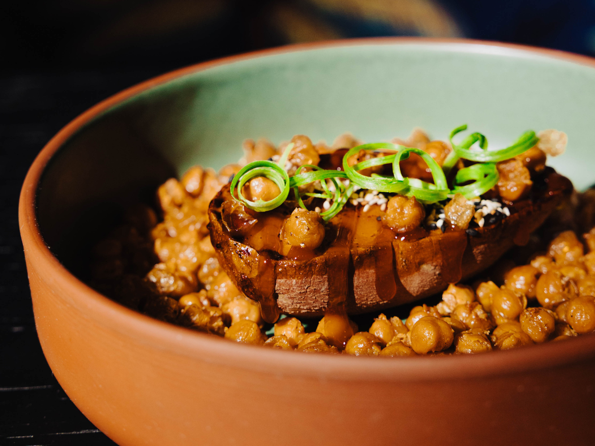 A bowl of roasted chickpeas topped with a halved sweet potato, drizzled with sauce and garnished with sliced green onions.