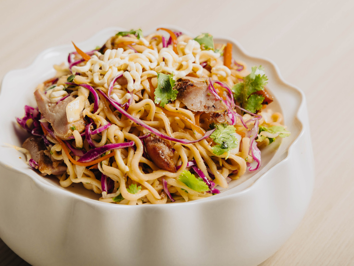 A white bowl filled with noodle salad, featuring shredded cabbage, sliced meat, carrots, cilantro, and a light dressing.