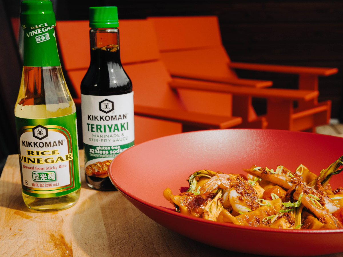 A bowl of stir-fried vegetables sits next to bottles of Kikkoman rice vinegar and teriyaki sauce, with orange patio chairs in the background.