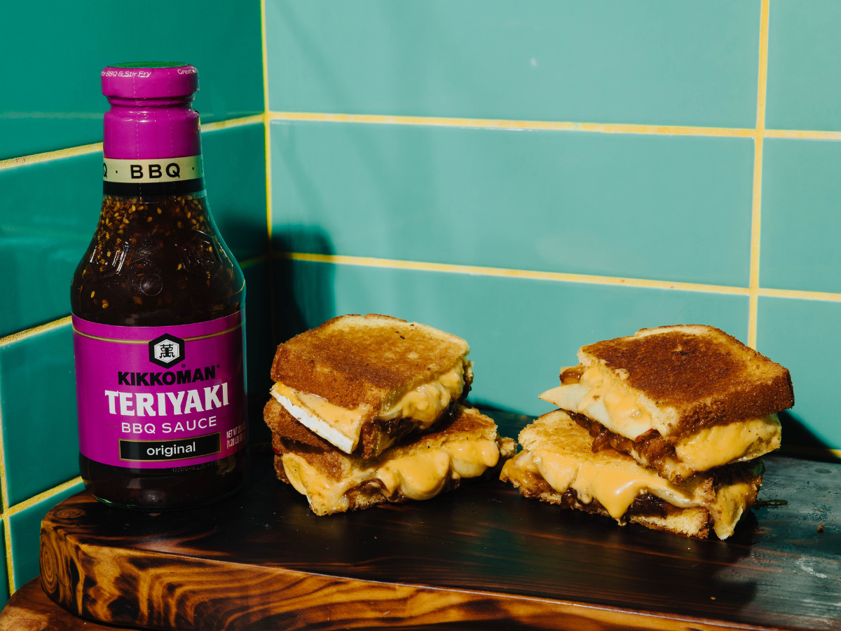 A bottle of Kikkoman Teriyaki BBQ sauce stands next to two grilled cheese sandwiches stacked on a wooden board, in front of teal tiled walls.