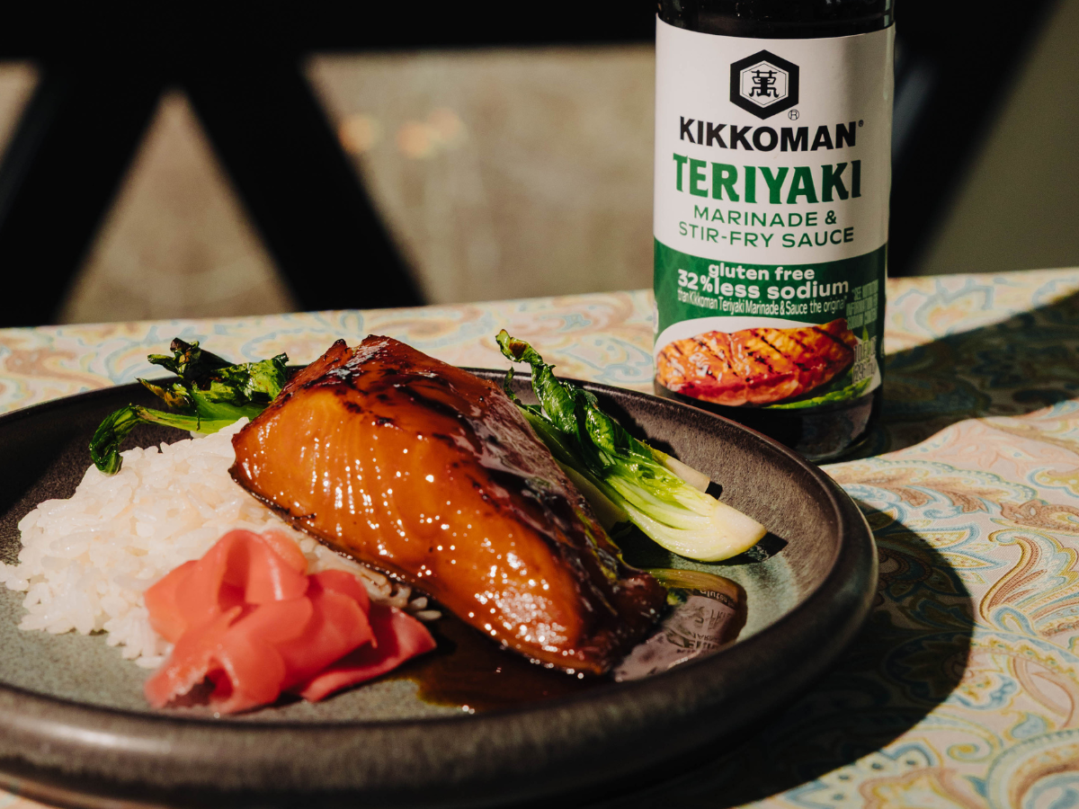 A plate with glazed fish, white rice, pickled ginger, and greens sits beside a bottle of Kikkoman Teriyaki Marinade & Stir-Fry Sauce.