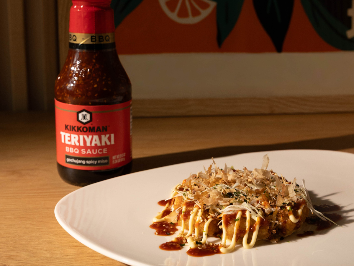 A white plate with okonomiyaki topped with sauces and bonito flakes sits beside a bottle of Kikkoman Teriyaki BBQ Sauce on a wooden table.