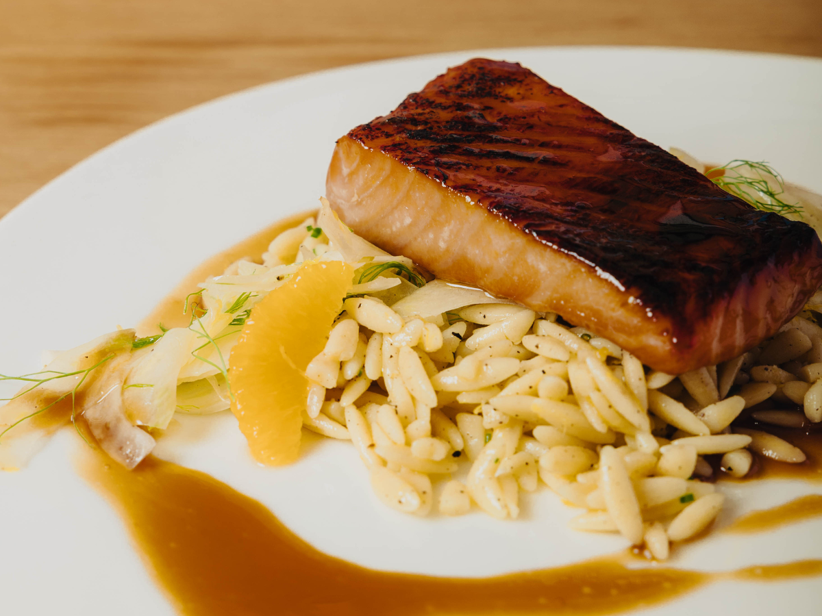 A glazed salmon fillet is served on a bed of orzo pasta with orange slices, fennel, and a brown sauce, presented on a white plate.