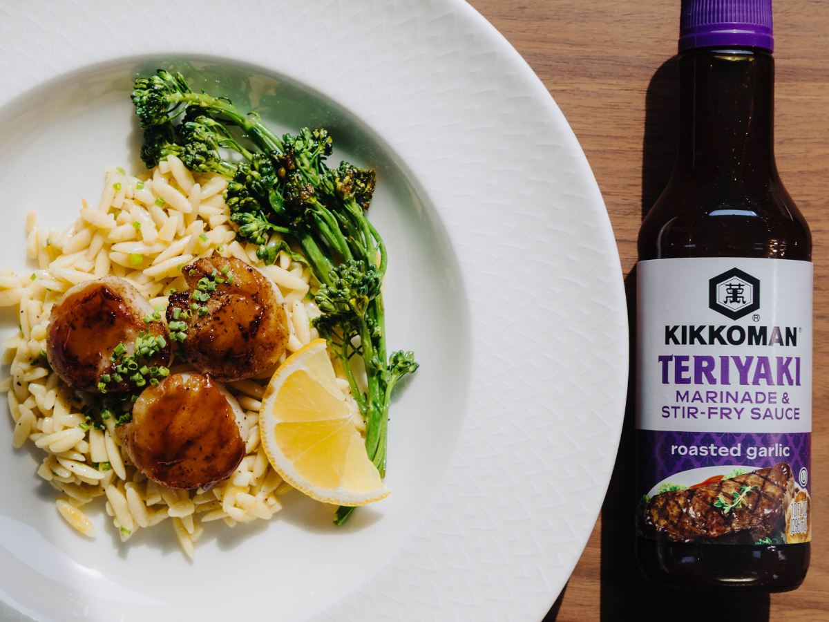 A plate with orzo, broccolini, three glazed scallops, and a lemon wedge next to a bottle of Kikkoman Teriyaki Marinade & Stir-Fry Sauce on a wooden surface.