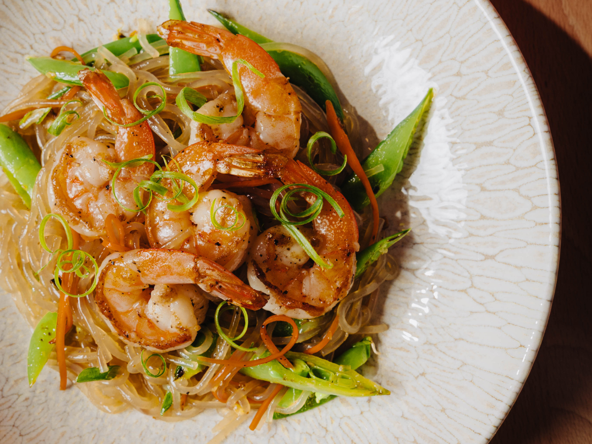A plate of shrimp stir-fry with glass noodles, sliced snap peas, carrots, and green onions, served on a textured white dish.