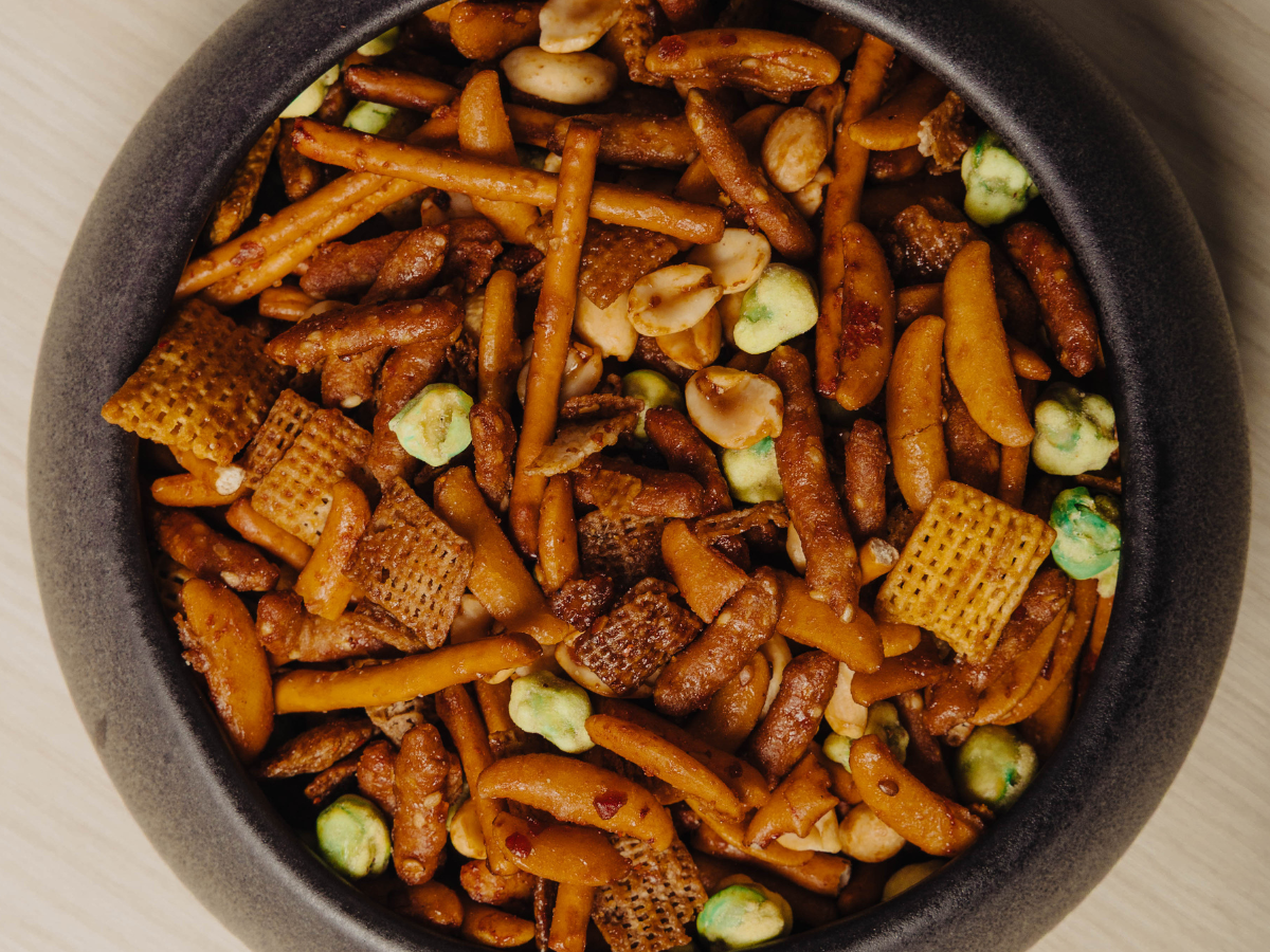 A bowl filled with a snack mix containing pretzel sticks, cereal squares, peanuts, rice crackers, and green wasabi peas.