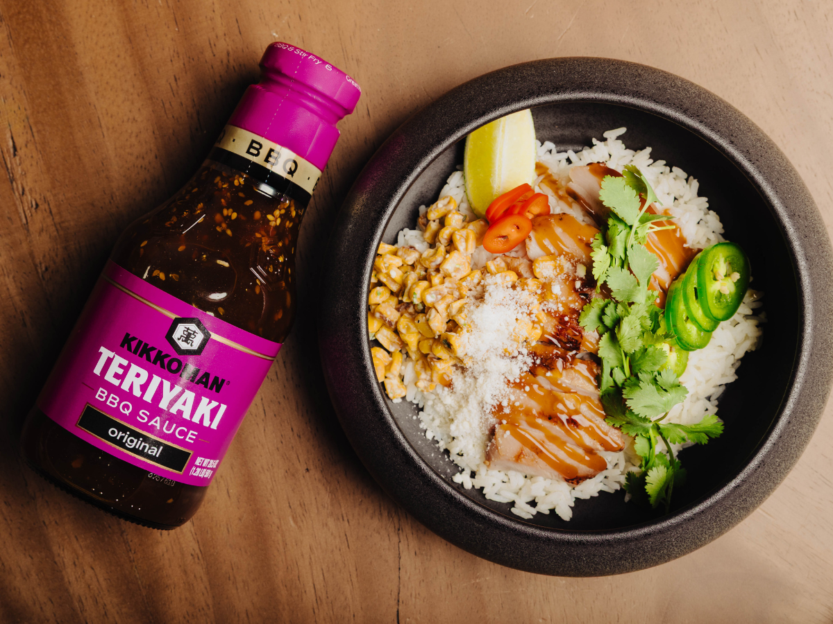 A bottle of Kikkoman Teriyaki BBQ Sauce next to a bowl of rice topped with sliced chicken, corn, cilantro, sliced peppers, a lemon wedge, and shredded cheese.