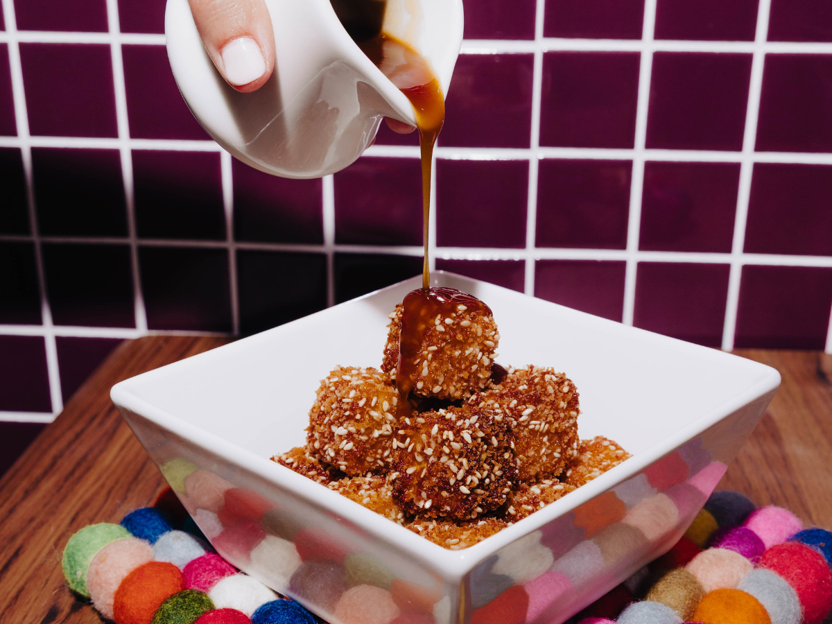 A hand pours sauce over breaded food pieces in a white square bowl on a colorful felt mat with a purple tiled wall in the background.
