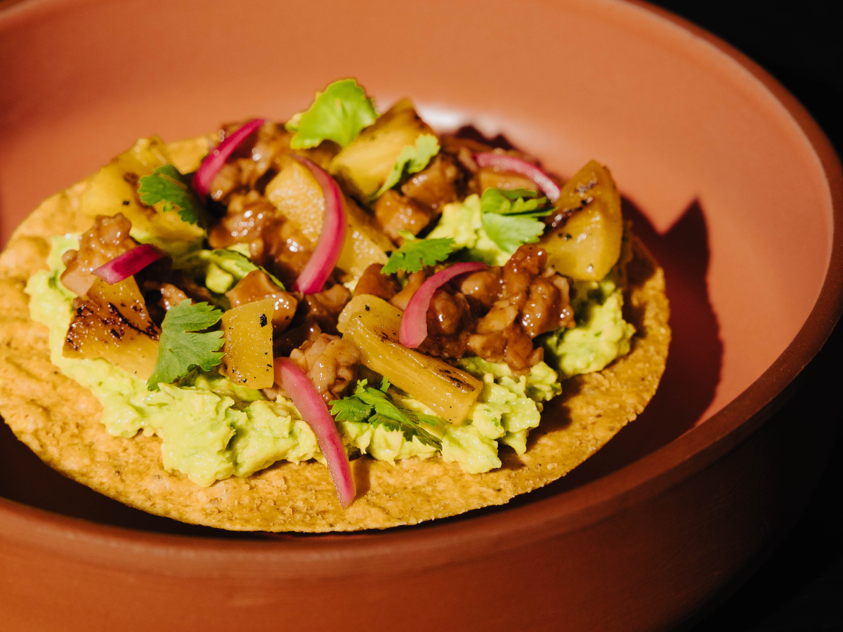 A tostada topped with mashed avocado, grilled pineapple, marinated meat, red onion slices, and cilantro sits in a brown bowl.