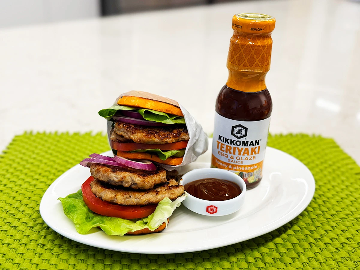 Two turkey burgers with lettuce, tomato, and red onion are served on a white plate with a dish of sauce and a bottle of Kikkoman Teriyaki BBQ & Glaze on a green placemat.