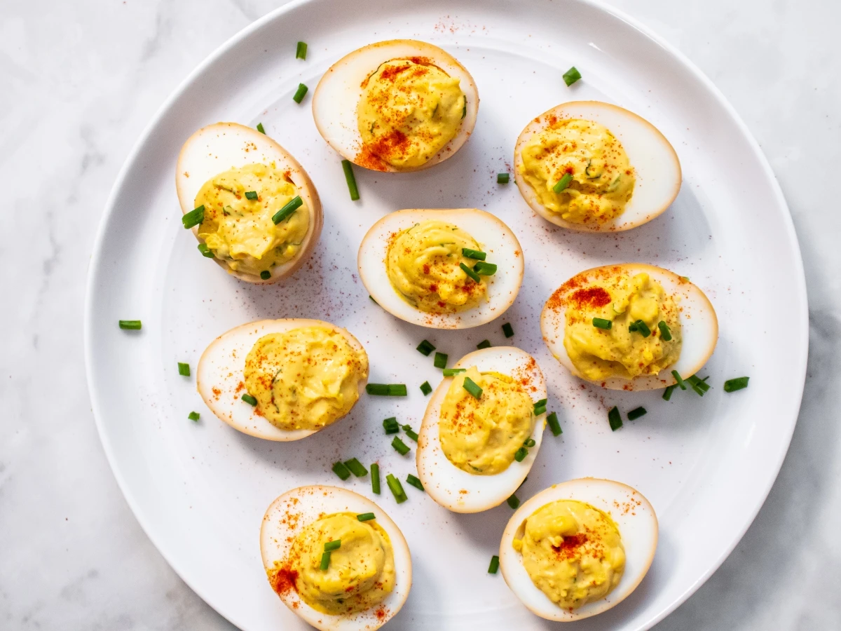 A white plate with eight deviled eggs, garnished with chopped chives and sprinkled paprika, on a marble surface.