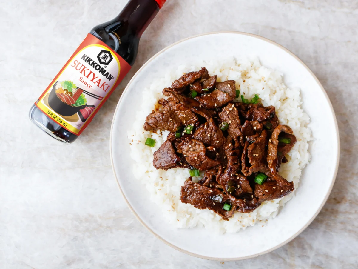 A plate of white rice topped with cooked beef and green onions sits next to a bottle of Kikkoman Sukiyaki Sauce on a marble surface.
