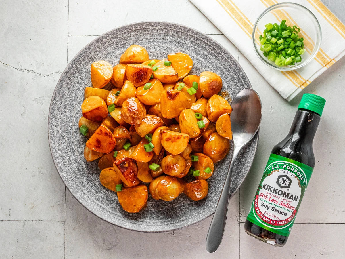 A plate of glazed, roasted potatoes garnished with chopped green onions sits next to a spoon, a small bowl of green onions, and a bottle of Kikkoman less sodium soy sauce.