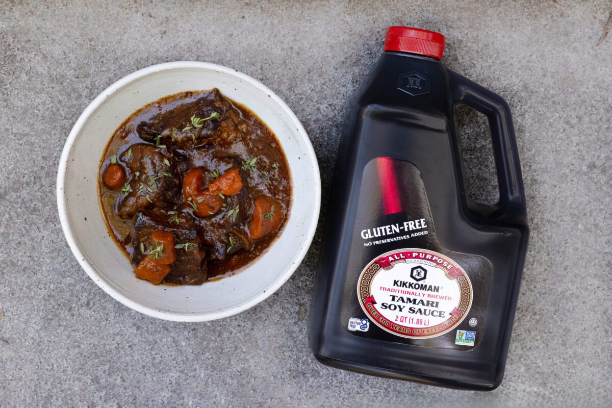 A white bowl of beef stew with carrots and herbs sits next to a large bottle of gluten-free Kikkoman soy sauce on a gray surface.