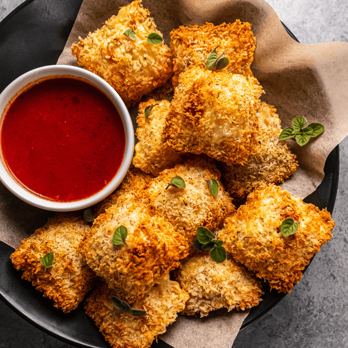 Golden, breaded ravioli served on parchment paper with a small bowl of red marinara sauce, garnished with fresh green herbs.