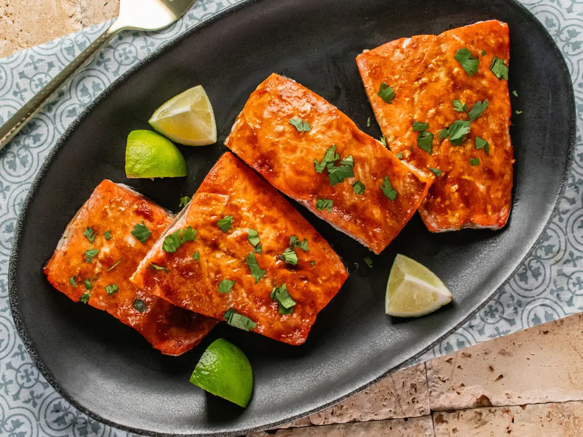 Four pieces of cooked salmon garnished with chopped herbs are arranged on a black oval plate, with lime wedges placed beside the fish.