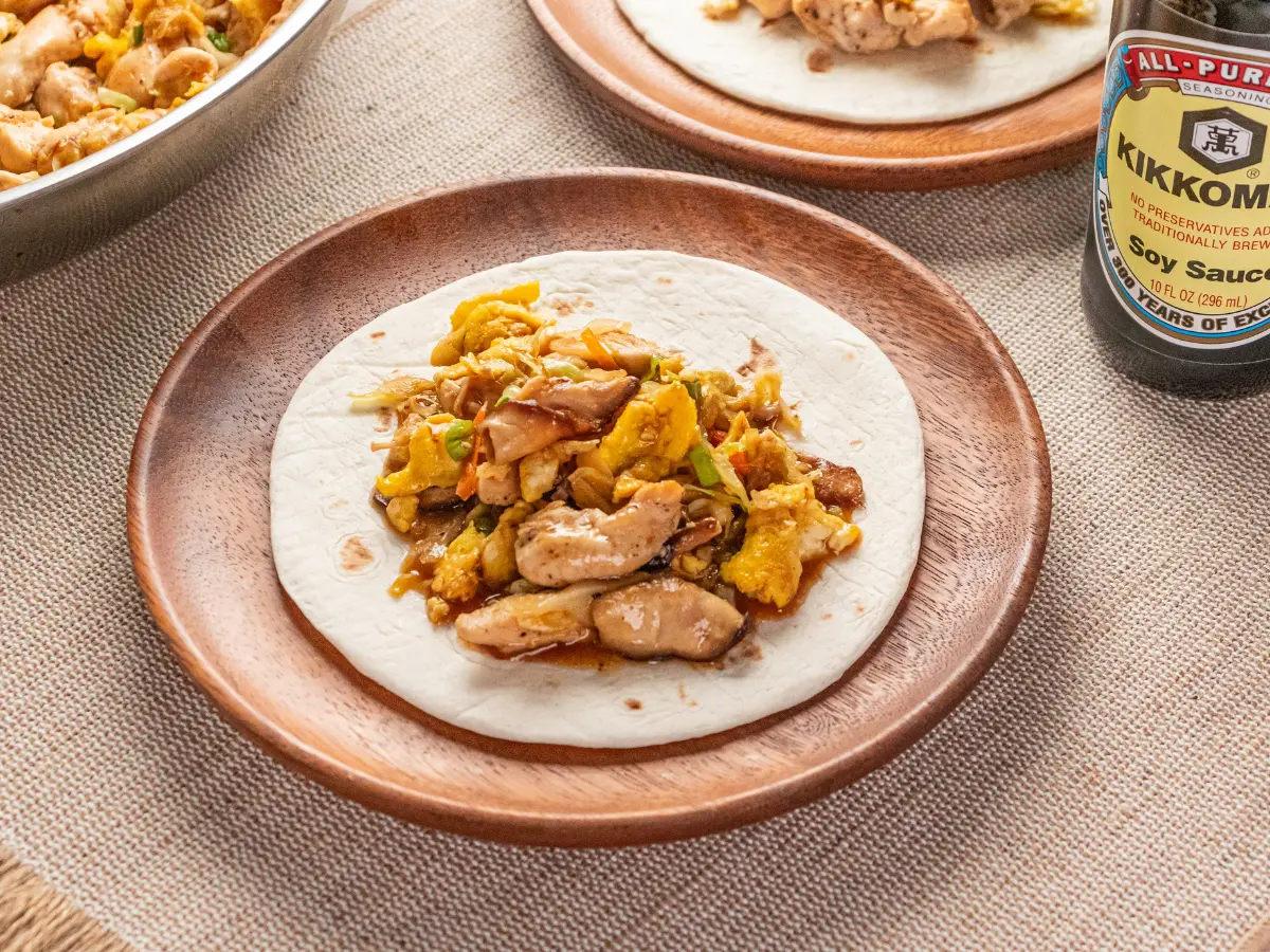 Flour tortilla topped with scrambled eggs, cooked chicken, and vegetables on a wooden plate; soy sauce bottle visible on the side.