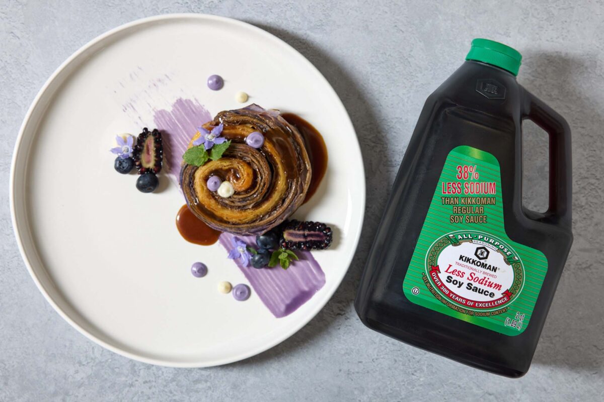 A plated pastry dessert with sauce and berries sits beside a jug of Kikkoman Less Sodium Soy Sauce on a gray surface.