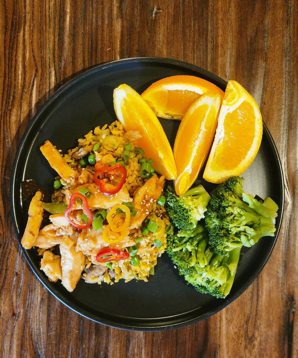 A black plate with broccoli, orange slices, and fried rice with chicken, peas, chopped green onions, and sliced red and yellow peppers on a wooden table.