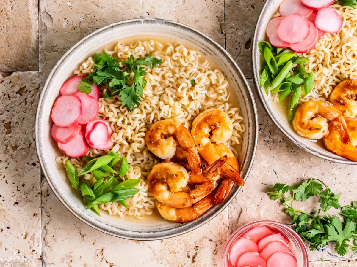 A bowl of ramen noodles topped with shrimp, sliced green onions, pickled radish, and cilantro sits on a tile surface next to another partially visible bowl.