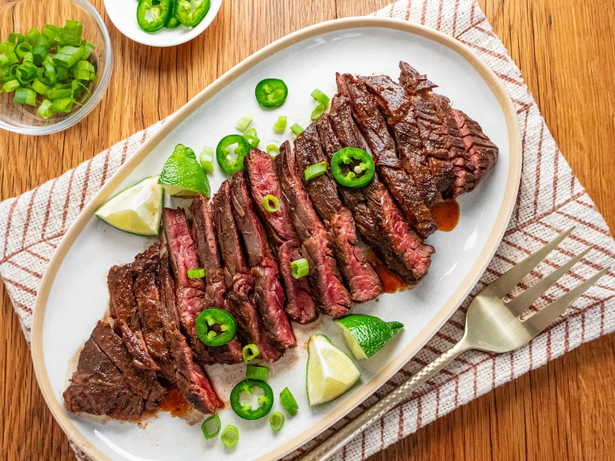 Sliced grilled steak garnished with jalapeño, green onions, and lime wedges on a white oval plate, placed on a cloth napkin with a fork nearby.