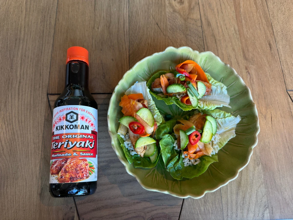 A bottle of Kikkoman Teriyaki sauce next to a green bowl containing three lettuce wraps with vegetables and red chili slices, on a wooden surface.