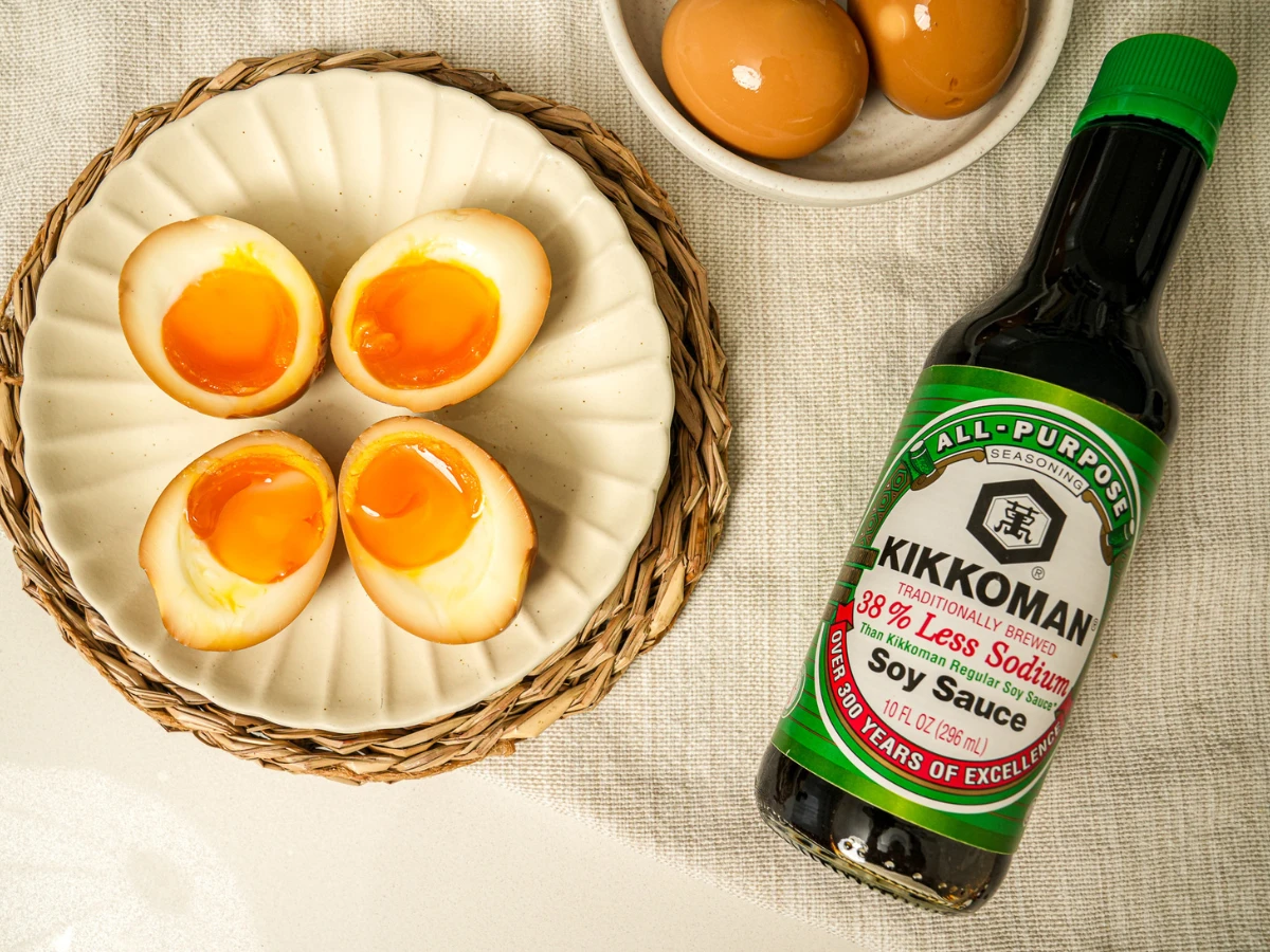Four halved soft-boiled eggs on a plate next to a bottle of Kikkoman low-sodium soy sauce and a bowl of whole marinated eggs.
