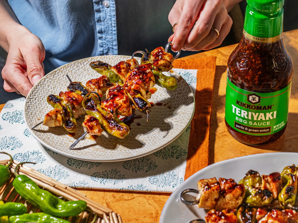 Person placing grilled chicken and vegetable skewers on a plate next to a bottle of Kikkoman Teriyaki BBQ Sauce.