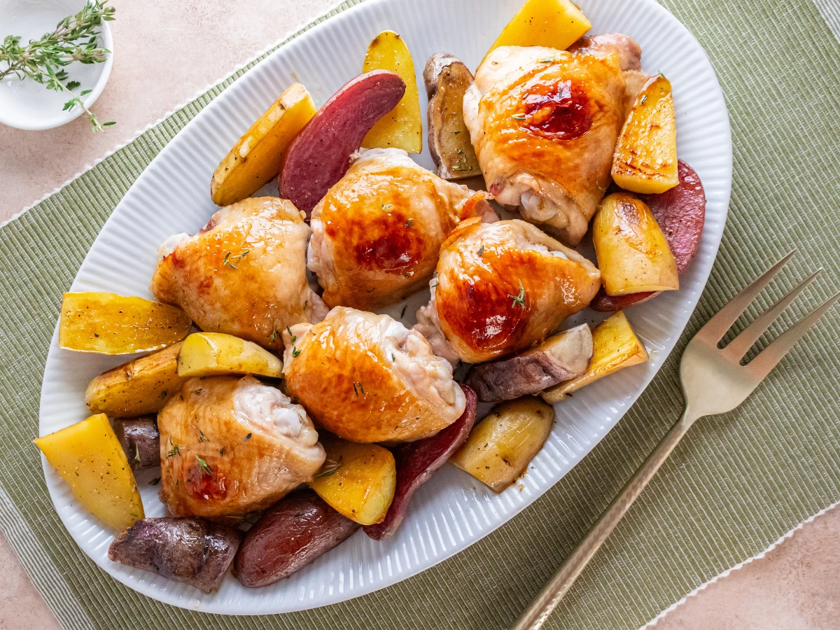 Roasted chicken thighs and assorted potatoes are arranged on a white oval platter with a gold fork beside it.