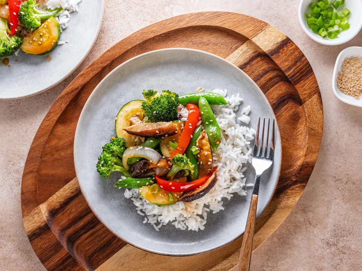 A plate of white rice topped with stir-fried vegetables including broccoli, snap peas, zucchini, red pepper, and onion, with a fork on the side.