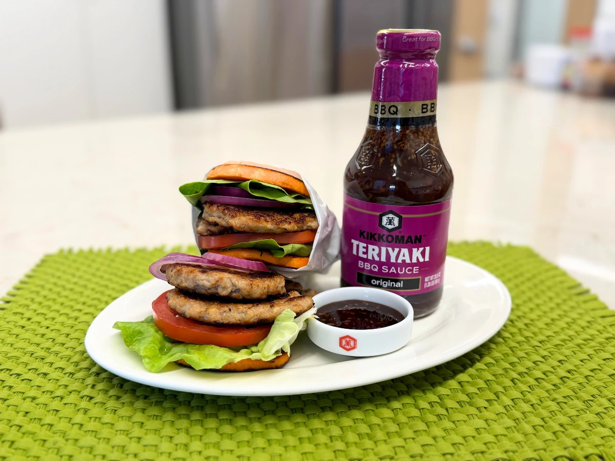 Plate with a stacked burger containing lettuce, tomato, onion, and patties, next to a bottle of Kikkoman Teriyaki BBQ sauce and a small dish of sauce on a green placemat.