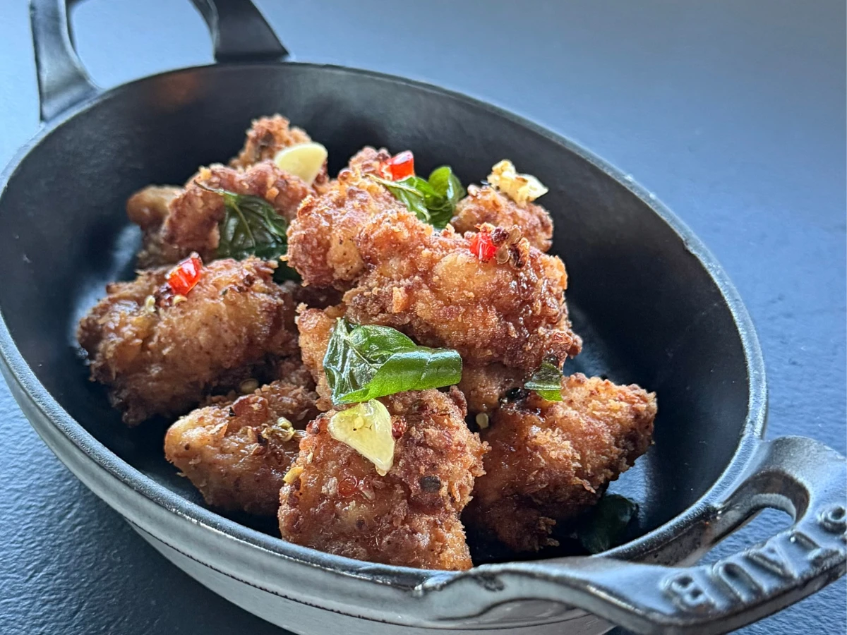 A Staub dish filled with pieces of fried chicken garnished with sliced garlic, red chili, and green herbs.