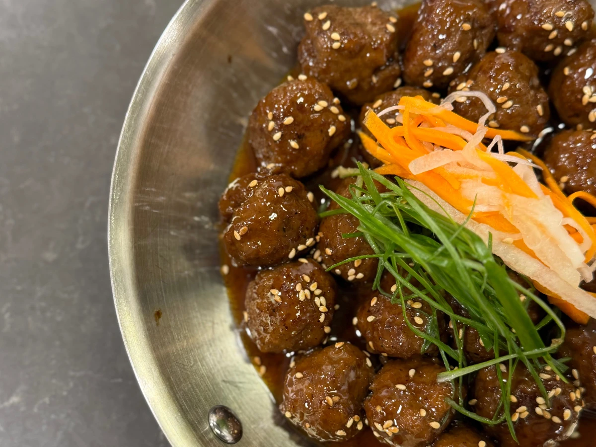 A metal bowl filled with glazed meatballs topped with sesame seeds, shredded carrots, daikon, and sliced green onions sits on a dark surface.