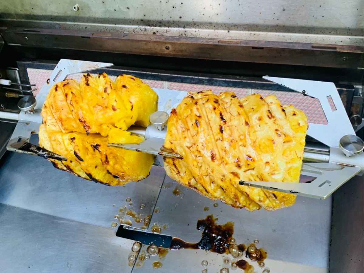 Two pineapples are skewered on a rotisserie inside a grill, with char marks and caramelized juice visible on the metal surface below.